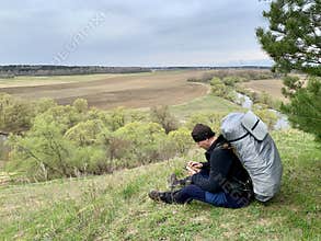 Narofominsky district, Moscow region, Russia, May, 02, 2021. Tourist with a cell phone sits on the high bank of the Protva River.