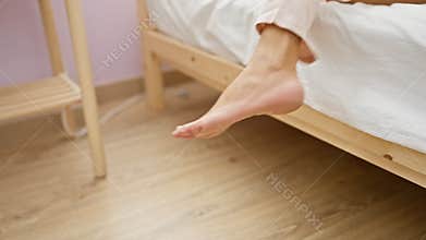 A peaceful scene capturing a woman\'s barefoot dangling from a wooden bed in a bedroom with light pink walls and minimalist
