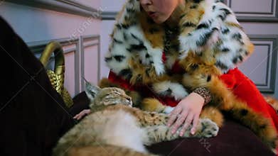 Woman in fur cloak blows on lynx calf which lies