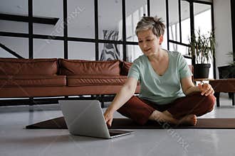 A middle aged lady is practicing yoga at home. A smiling woman sits on a mat on the floor in the lotus position, has a