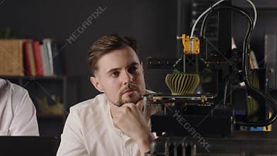 A concentrated young guy mechanical engineer with interest observes the working process of 3d printer. A digital machine