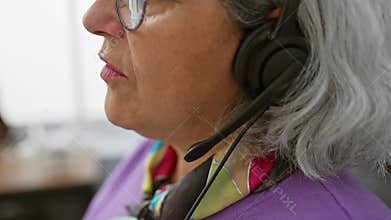 Professional senior woman with headset in modern office interior, glasses, grey hair, speaking, focused, business, support