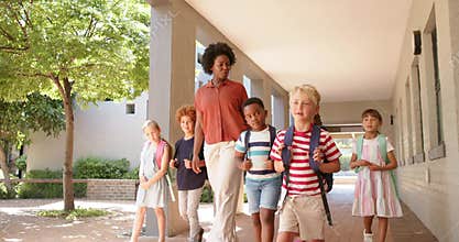 Teacher with children guiding along walkway with columns backpacks as blonde boy stepping to lead
