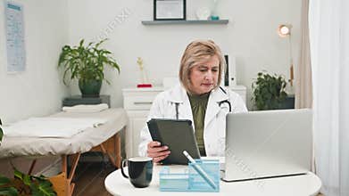 Mature Female Doctor Conducting Online Doctor Consultation with Tablet and Laptop in Modern Clinic Office