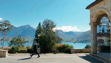Man in suit walks along scenic lakeside promenade with mountains and architecture backdrop view