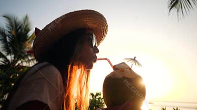Attractive Young Tourist Hipster Girl Drinking Fresh Thai Coconut Water Coctail at Beach Against Beautiful Sunset. 4K