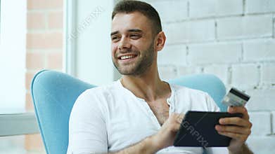 Young smiling man doing online shopping using digital tablet computer sitting at balcony at home