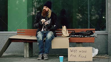 Homeless and jobless european man with cardboard sign eat sandwich on bench at city street because of immigrants crisis