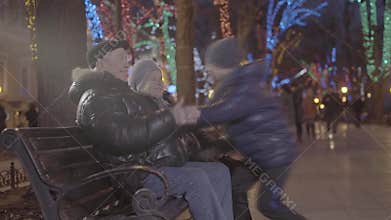 Excited happy grandchild boy running over to grandparents sitting on bench in christmas festive atmosphere evening park