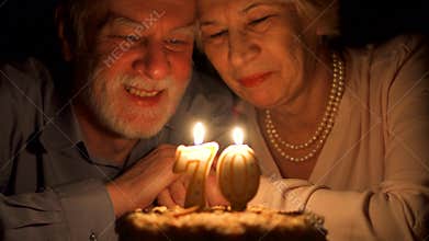 Loving senior couple celebrating anniversary with cake at home in the evening. Blowing out candles.