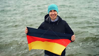 Slow motion portrait of joyful German sports fan holding flag of Germany and smiling while standing near the lake and