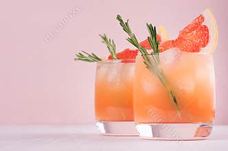 Two elegance cold wet drinking glasses with fresh grapefruit summer alcohol cocktail, ice, rosemary on fashion pink background.