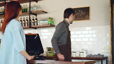 Attractive young woman is talking to friendly male cashier, paying for takeaway coffee with mobile phone at coffee shop