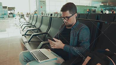 Male passenger sitting in waiting hall in airport and texting sms by smartphone