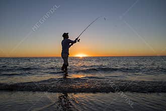 Silhouette of fisherman with hat on the beach with fish rod standing on sea water fishing at sunset with beautiful orange sky in v
