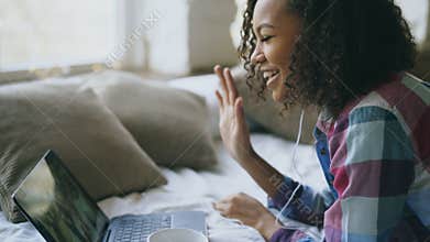 Curly african american young woman having video chat with friends using laptop camera while lying on bed