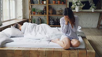 Sad woman is sitting on bed and crying after fight with her boyfriend while he is lying in bed with his back to her