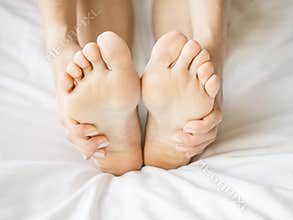 Woman Holding her Tired Feet in Hand Sitting on Bed with White Sheets