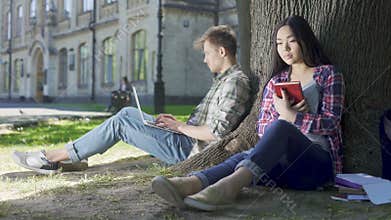 Multinational girl sitting under tree, holding book, looking at guy, feelings