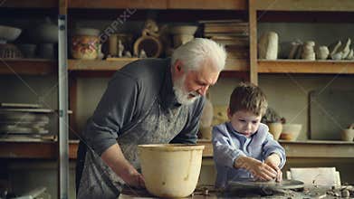 Pottery teacher senior adult is helping young student to form pot from piece of clay on throwing-wheel. Cute boy is
