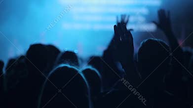 People raising up hands at a concert. A close-up. A back view. Singing. Blurred stage.