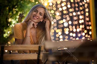 Smiling blonde young woman sitting, with evening fairy lights on the background.