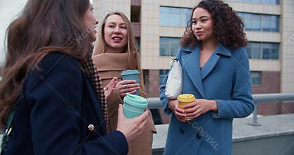 Togetherness concept. Beautiful multiethnic young women friends talk outside with coffee in eco cups on cold cloudy day.