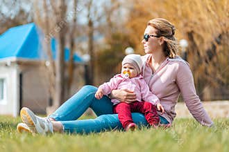 A happy mother sits on the grass, hugging her baby child, enjoying the spring sun. Family outdoor recreation with children