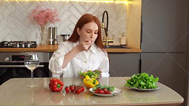 Attractive young redhead woman cutting fresh cucumber cooking food salad sitting at table in modern kitchen room.