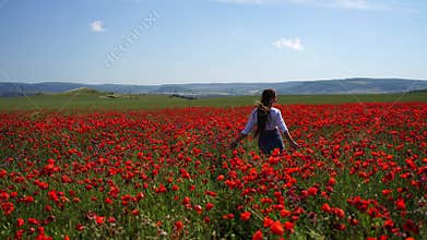 A beautiful young woman with long hair runs on a poppy field. She is wearing a white shirt and denim skirt.