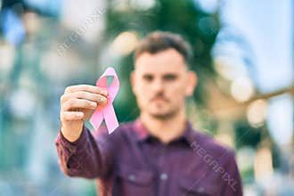 Young hispanic man with serious expression holding pink breast cancer ribbon at the city