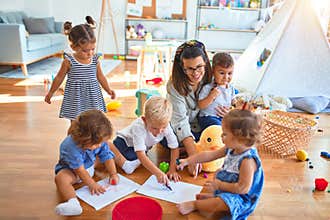 Beautiful teacher and group of toddlers sitting on the floor drawing using paper and pencil around lots of toys at kindergarten