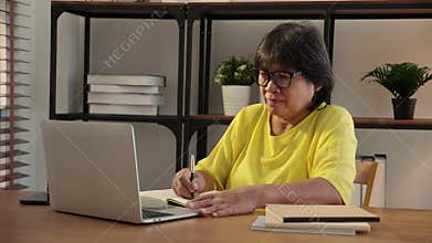 Senior asian woman working with writing on note and study while laptop on desk office in living room.