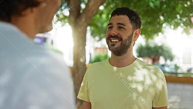 Two men laughing and chatting together in a sunny urban park with trees and buildings in the background, highlighting their