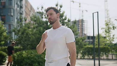 A young sportive man does morning exercises, stretches arms and neck before jog in the city park area. An athlete guy