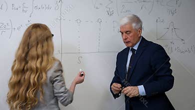 An elderly professor explains a subject to a student at a white board.