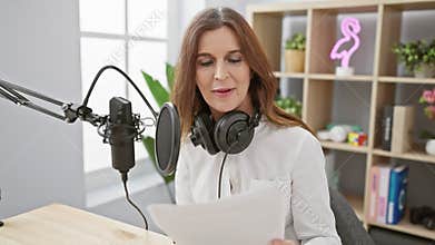 A middle-aged woman in a white shirt podcasting in a modern studio with a microphone and headphones