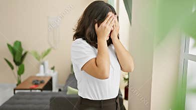 Shy, beautiful young hispanic woman peeking through her fingers as shock covers her face at home, while expressing a mix of fear