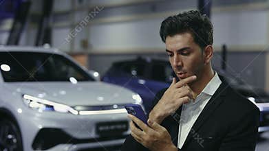 Handsome Male Consultant Browsing on Smartphone, Texting with Potential Clients Near Automobile in Dealership