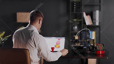 A man graphic designer sits by the table, uses calipers and measures the parameters of red plastic funnel. A sketch of