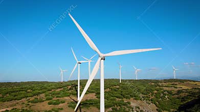 erial drone footage of a wind farm with large wind turbines spinning over green fields under a clear blue sky. The video