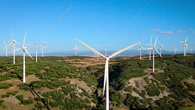 erial drone footage of a wind farm with large wind turbines spinning over green fields under a clear blue sky. The video