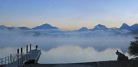 At dawn, Lake McDonald in Glacier National Park reflects mountains and fog in the calm water: panorama