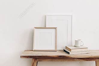 Wooden square and white portrait frame mockups on vintage bench, table. Cup of coffee on pile of books. White wall