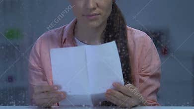 Upset lonely lady sitting behind rainy window and reading letter, missing family