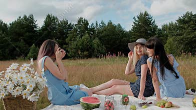 Three young attractive girls on a picnic. The photographer takes pictures on the mirrorless camera of two models. Models