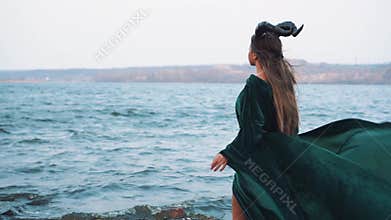 Mystic lady with long hair in flying gorgeous green dress, witch in emerald velor gown on shore of magical lake, girl