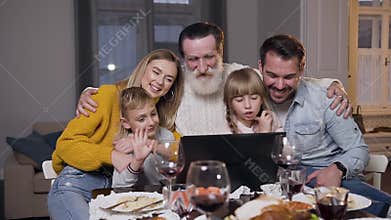 Attractive different generation family having video chat on computer during festive dinner and greeting their