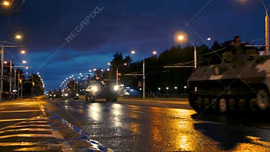 A convoy of military equipment rides through the city at night with headlights on