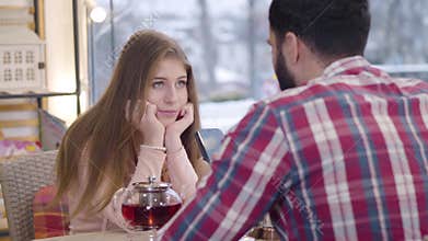 Portrait of bored pretty Caucasian girl sitting with boyfriend or husband in restaurant as her spouse using smartphone